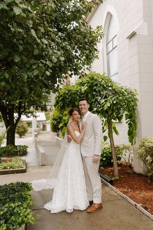 Smiling bride and groom pose together under green trees beside a white church on their wedding day.