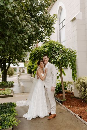 Bride in lace gown and groom in tan suit embrace warmly under trees outside a white wedding church.