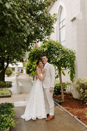 Smiling bride and groom embrace beside a lush green tree outside a white church on their wedding day.