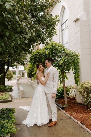 Bride in lace gown and groom in tan suit pose near a vine-covered white building.