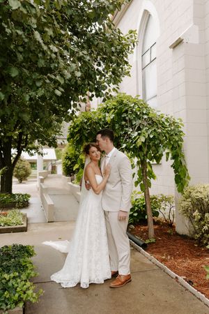 Groom in tan suit embraces bride in white lace gown beside a white church with climbing vines.