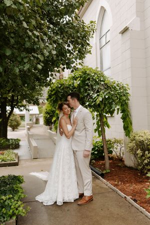 Bride and groom share a romantic moment under lush green trees outside a white stone church.