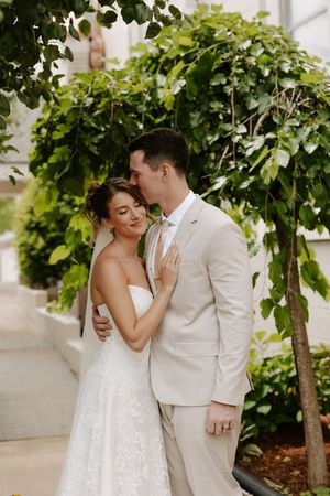 Groom in beige suit kisses bride on the cheek as she smiles softly beneath a canopy of green leaves.
