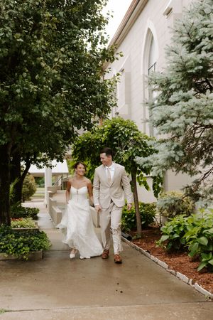 Bride and groom walk hand-in-hand along a garden path outside a white church on their wedding day.