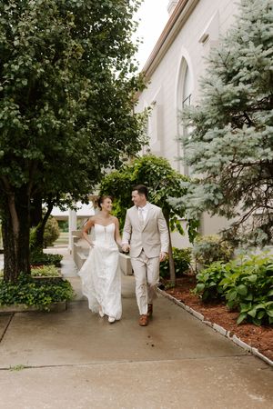 Bride and groom walking hand in hand along garden path beside historic church building on wedding day.