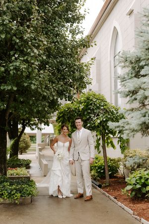 Bride and groom pose hand in hand on a garden path beside a white stone church.