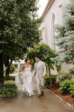 Smiling bride and groom in tan suit walking hand in hand along garden path beside church.