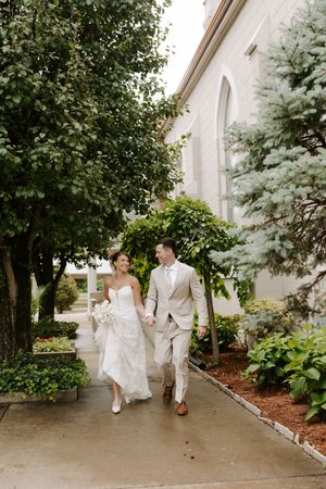 Happy bride and groom walk hand in hand along a garden path outside an elegant white church.