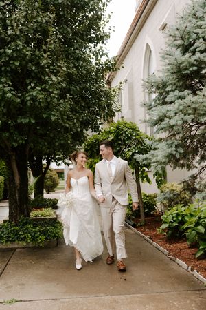 Bride and groom walk hand-in-hand smiling along pathway beside white church and lush greenery.