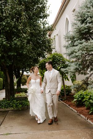 Bride and groom walk together smiling along a garden path beside a white stone building.