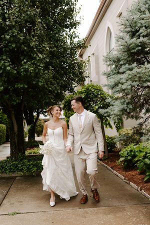Bride and groom walk hand in hand laughing together along a garden path outside a white church.