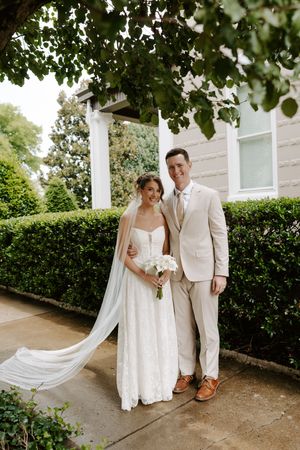 Smiling bride in lace gown with veil and groom in tan suit pose together outdoors on wedding day.