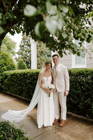 Bride and groom posing together under leafy archway beside manicured hedges, she holds white bouquet, veil flowing.