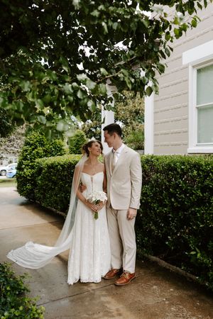 Smiling bride and groom standing together on a garden path beside trimmed hedges, veil flowing in the breeze.