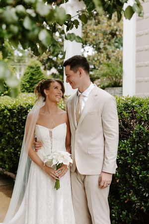 Bride and groom gaze at each other under leafy branches near white columns, she holds calla lilies in a lace bridal gown.