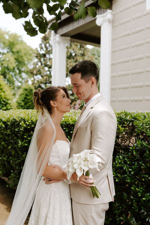 Bride and groom share a tender moment outdoors, she in a lace gown with veil, he in a beige suit holding calla lilies.
