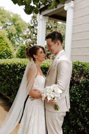 Bride and groom embrace near white columns and hedges, she wears a lace gown with veil, he wears a beige suit.