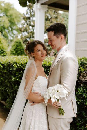 Bride in lace gown with veil leans into groom holding white calla lily bouquet beside a white column outdoors.