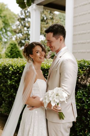 Bride smiling at groom beside white columns and hedges, he holds white calla lily bouquet, both in wedding attire.
