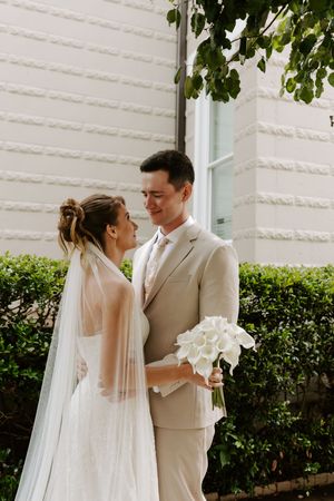 Bride and groom gazing at each other tenderly outside white building, groom holding white calla lily bouquet.