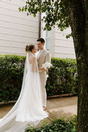Bride and groom share a romantic moment outdoors, bride in white gown with veil, groom in beige suit holding bouquet.