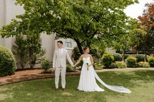 Newlyweds hold hands on a green lawn with a large tree and white building backdrop on their wedding day.