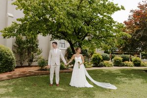Bride and groom hold hands walking across green lawn with large trees in background.
