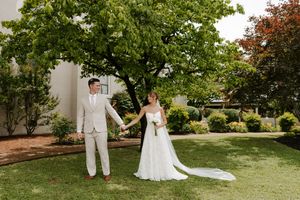 Bride and groom hold hands walking across green lawn with large trees at outdoor wedding venue.