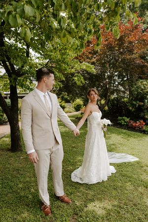 Bride and groom holding hands in lush garden, groom in beige suit leading bride in lace gown with veil.