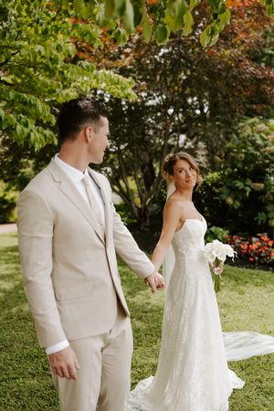 Bride in lace gown holds groom's hand while looking back, surrounded by colorful trees on a green lawn.