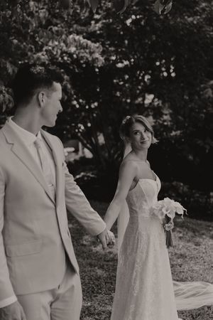Black and white photo of a bride and groom holding hands while walking through a lush garden on their wedding day.