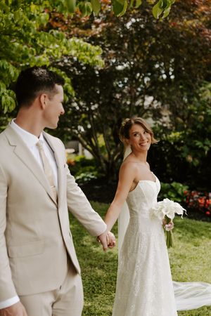 Bride in lace gown looks back smiling while holding grooms hand in sunlit garden.