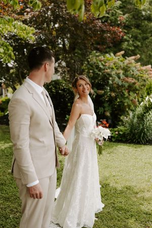 Bride in white lace gown holds bouquet while holding hands with groom in beige suit in lush garden.