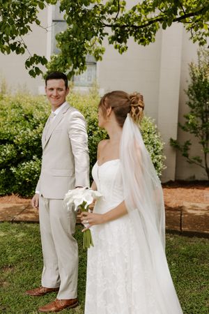 Bride and groom share a tender moment outdoors, groom in beige suit smiling back at bride in lace gown holding white calla lilies.