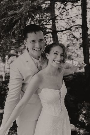Black and white photo of a smiling bride and groom embracing under pine trees on their wedding day.