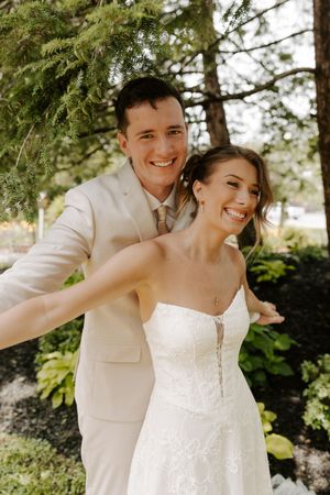 Joyful bride and groom embracing and laughing together outdoors under evergreen trees.