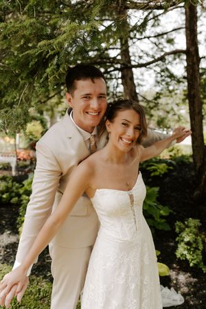 Joyful bride and groom posing together under evergreen trees, both smiling broadly in outdoor garden setting.