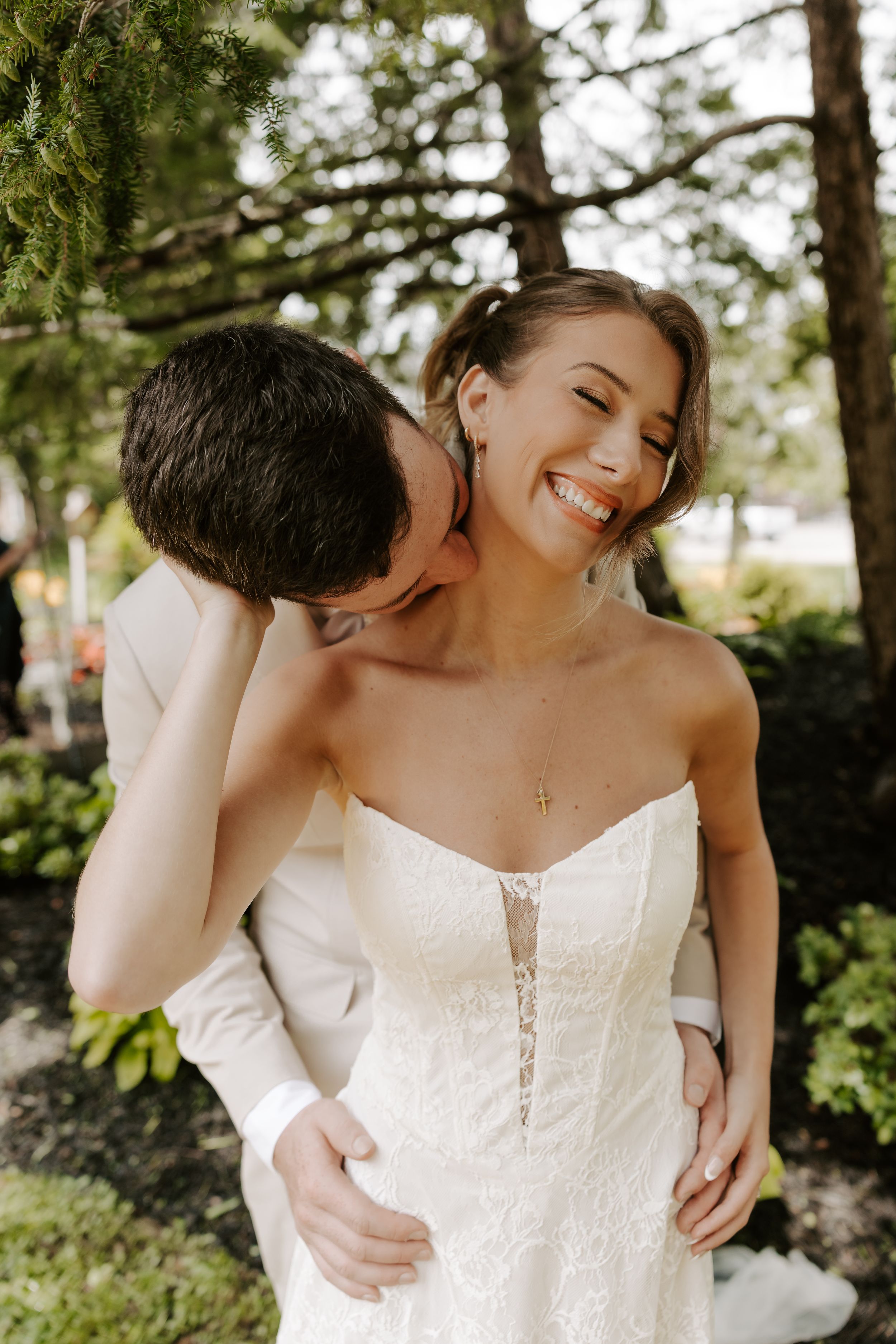 Groom kisses smiling bride on cheek as he embraces her from behind in lace strapless gown.