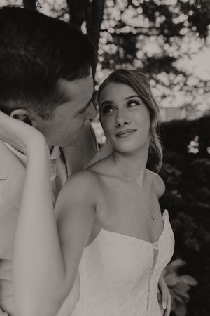 Black and white romantic portrait of bride in strapless gown gazing up at groom beneath garden trees.