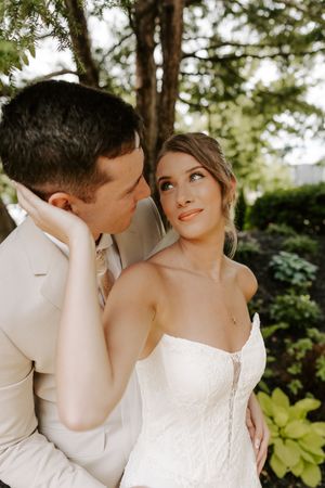 Bride in strapless lace gown gazes up at groom in beige suit as they embrace tenderly in a lush garden setting.