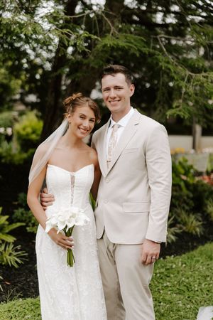 Bride in strapless lace gown holding calla lilies stands with groom in tan suit outdoors.