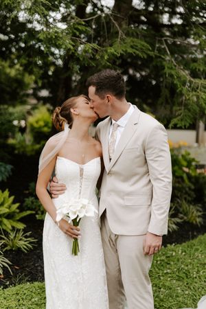 Bride and groom share a kiss outdoors, she holds white calla lilies bouquet, he wears a beige suit.