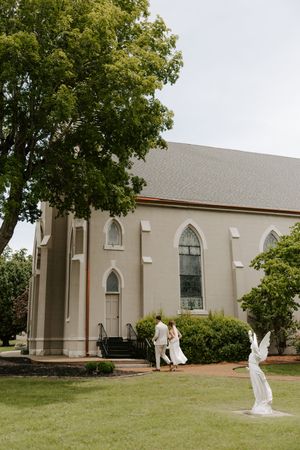 Bride and groom walking toward historic gray stone church with Gothic windows surrounded by green lawn.