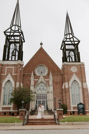 Bride and groom stand together on steps of a grand historic brick church with tall Gothic steeples.