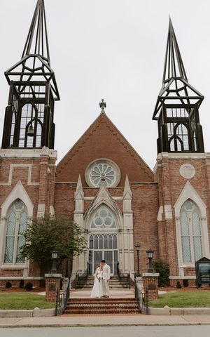 Bride and groom pose on steps before a grand Gothic brick church with twin spires on their wedding day.