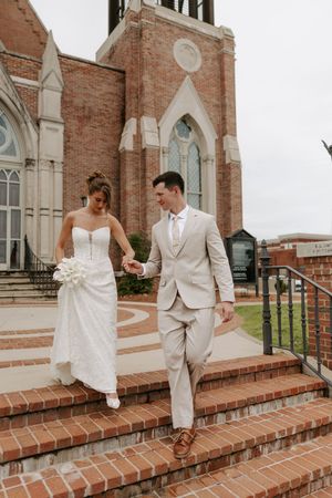 Bride and groom hold hands walking down brick church steps, she in white strapless gown, he in tan suit.