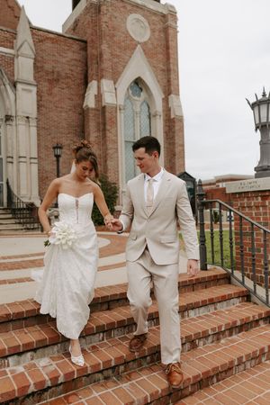 Bride and groom descend brick church steps hand-in-hand, bride in strapless gown and groom in tan suit.