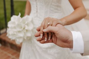 Close-up of bride and groom holding hands showing wedding ring with white floral bouquet in background.