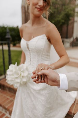 Close-up of bride and groom holding hands showing wedding rings and white bouquet.