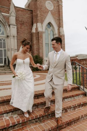 Bride and groom walk down brick church steps holding hands, she in lace gown, he in beige suit.